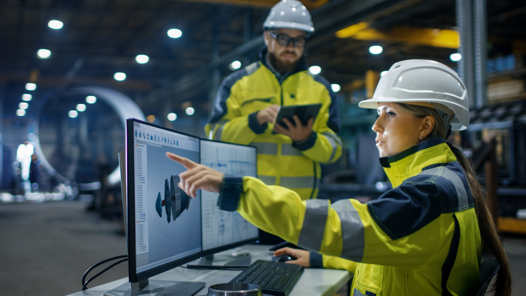 Two colleagues in yellow safety clothing and helmets work at a computer on a factory floor