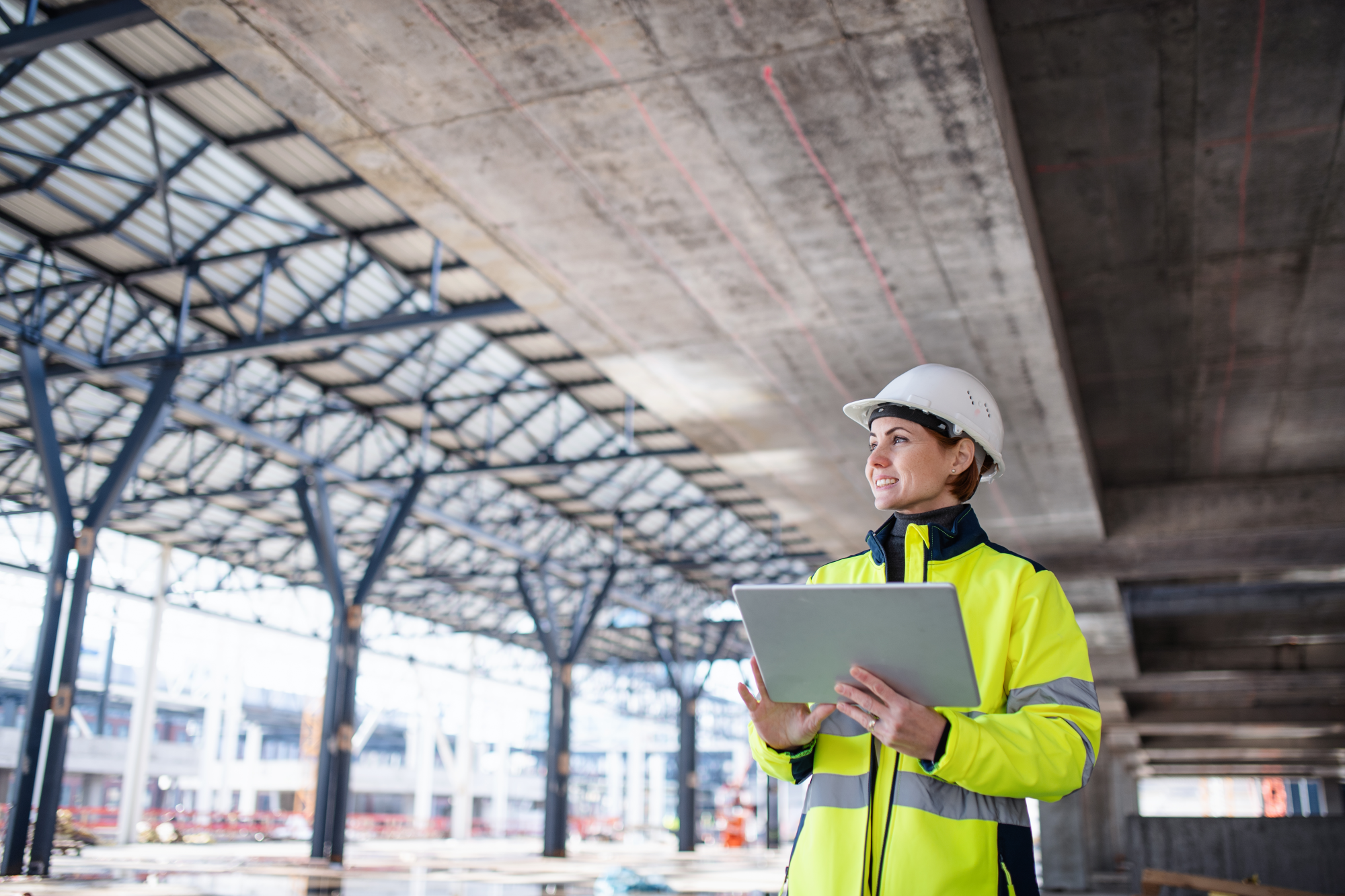 A woman in yellow work clothes and a white hard hat stands on a construction site and works on her t