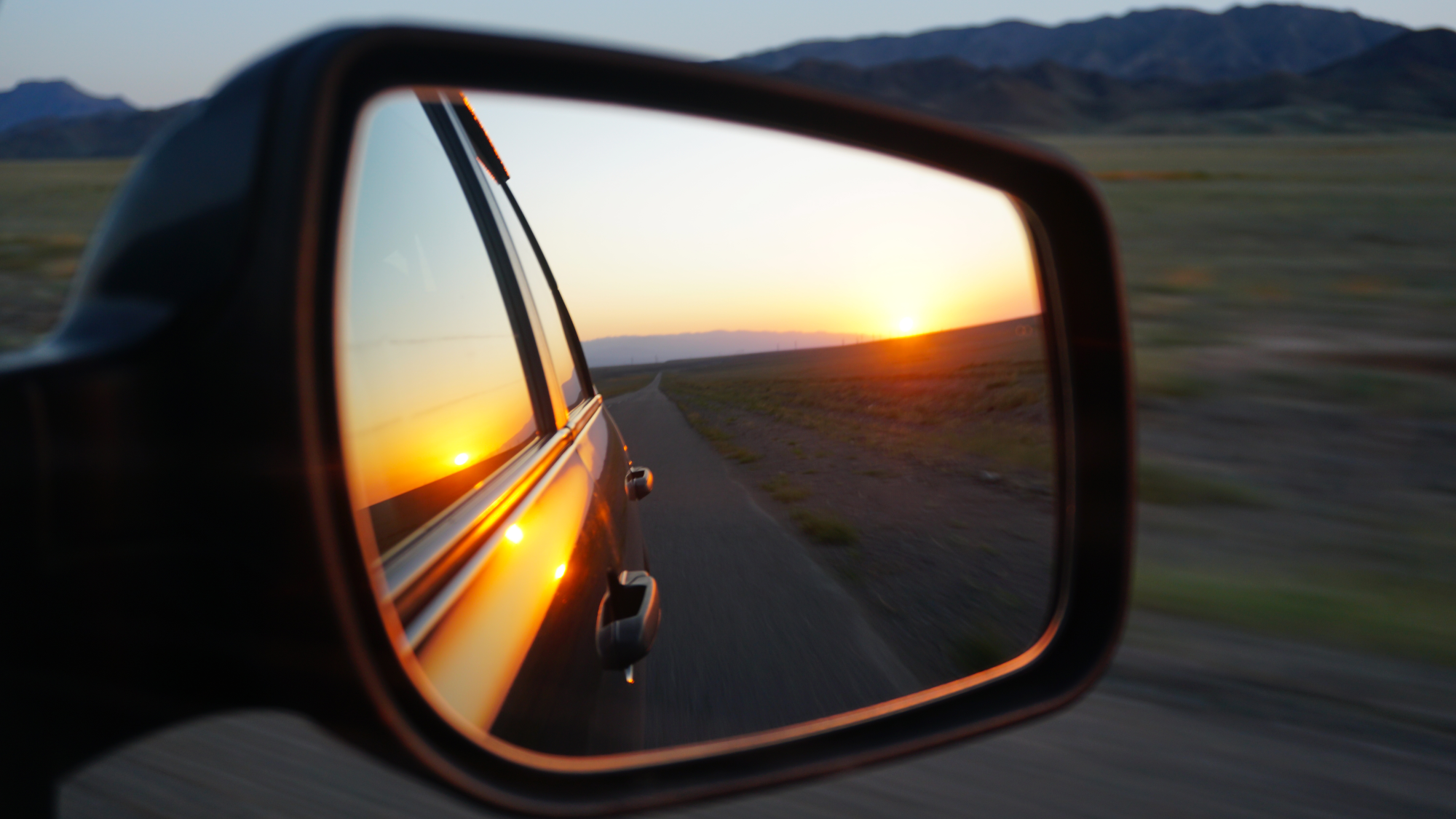 View in the side mirror of the car. Orange dawn over the hills. The car is going at high speed. Gree