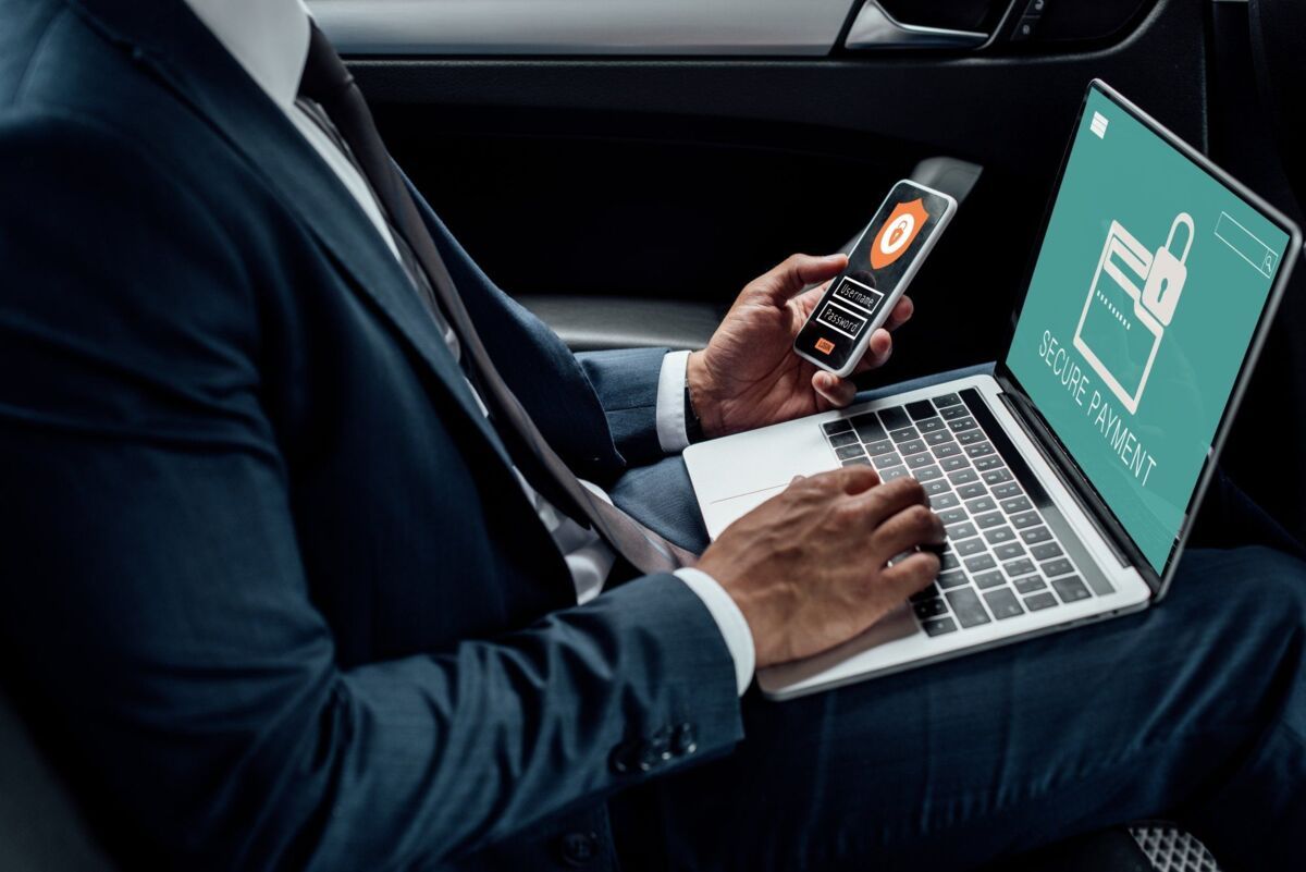 a man types on a laptop with one hand and a cell phone with the other while sitting in a car