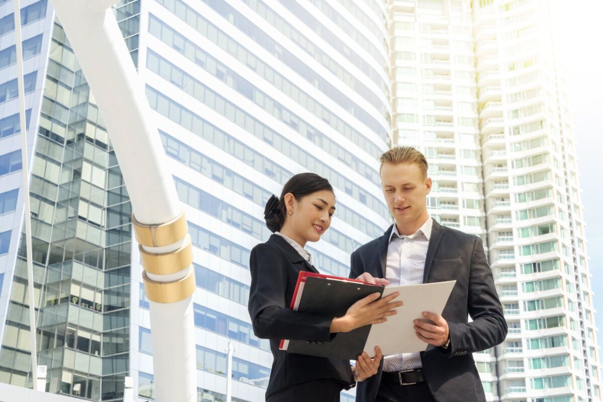 tcp-III-med-dqs-two businessmen look together in workbook near skyscrapers