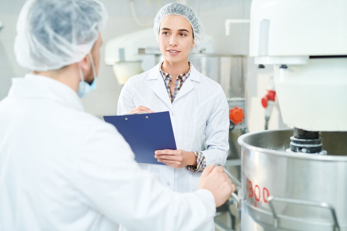 Confectionery factory employee standing near machinery in white coat making notes and speaking with 