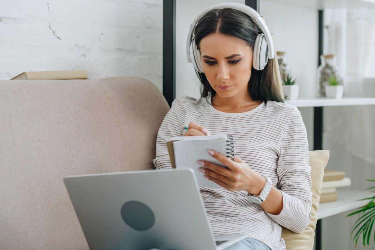 woman sits on the couch with a laptop and a headset taking notes on a notepad during a workshop