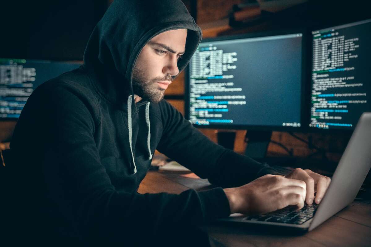 informationssicherheit-kmu-dqs-mann in pullover mit kapuze sitzt konzentriert am laptop umgeben von mehreren desktops