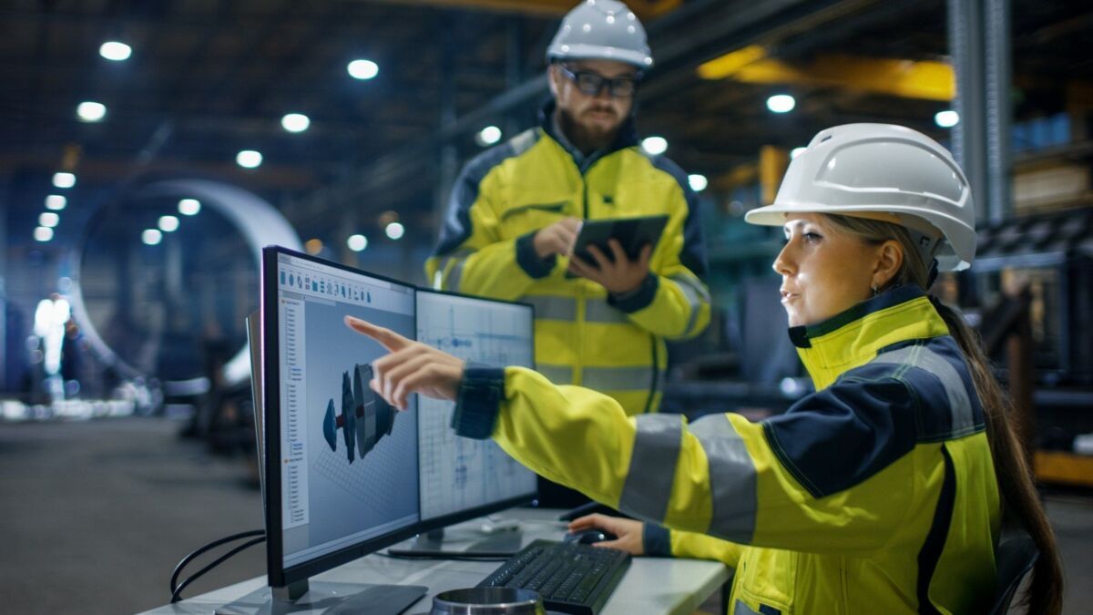 Two colleagues in yellow safety clothing and helmets work at a computer on a factory floor