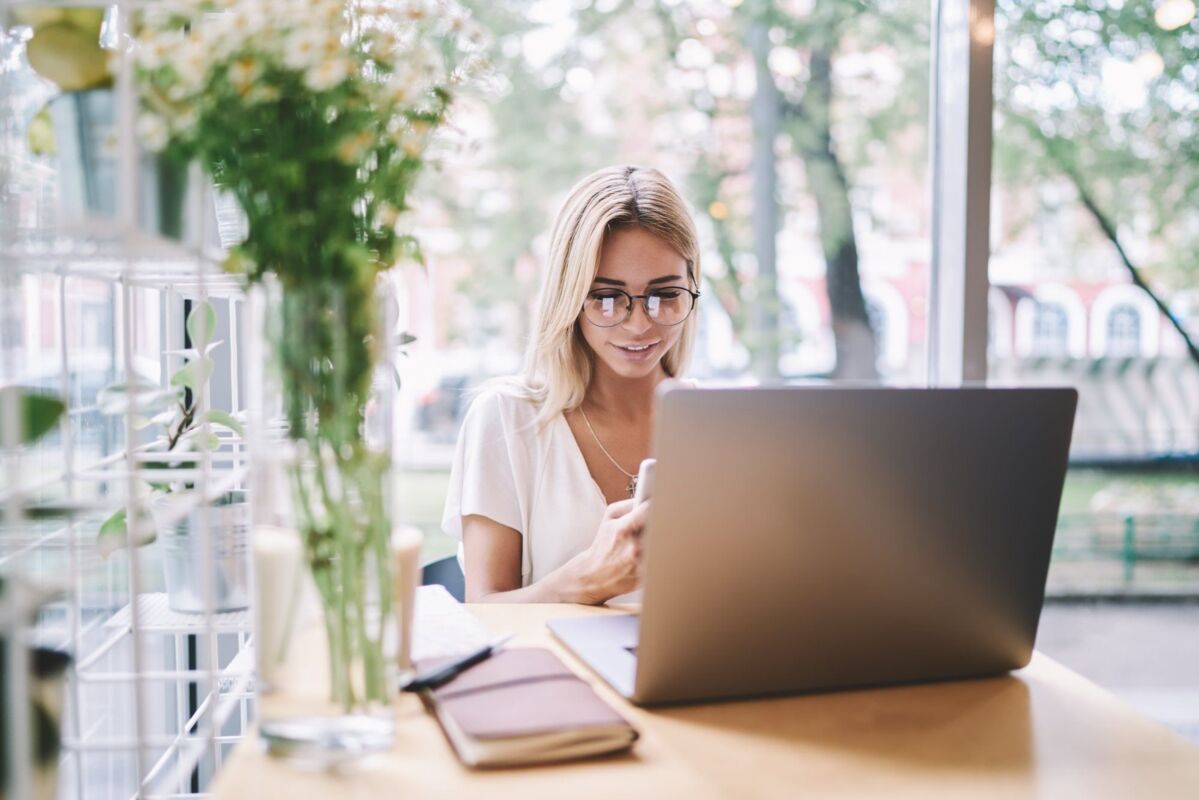 Smiling woman in eyewear updating notification on smartphone application for chatting while sitting 