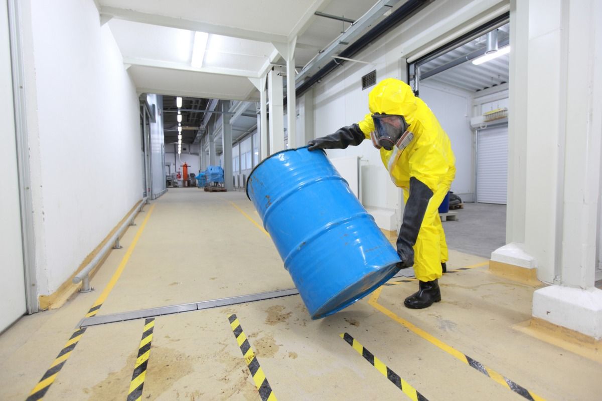 A person in a yellow full body protective suit rolling a light blue drum with hazardous waste down a long concrete corridor