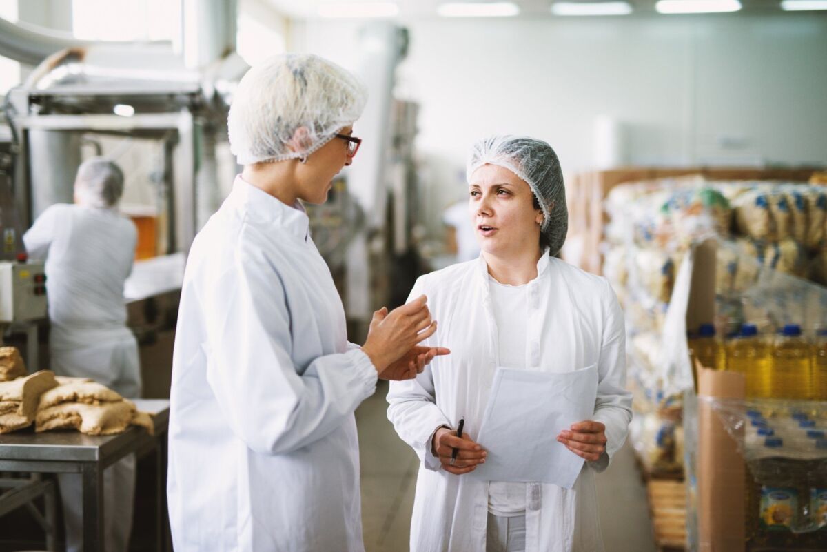 Two cheerful workers in white sterile cloths are standing in a food factory and talking while one of