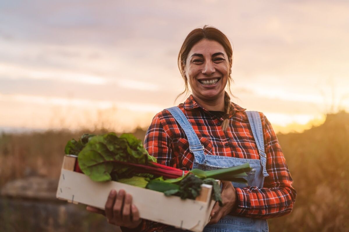 dqs-a smiling farmer stands in a field with a wooden box of fresh vegetables in her hand 