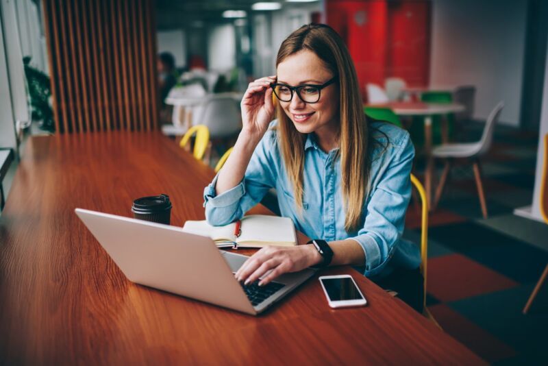 woman sits at a large table in a large, empty room and works on a laptop. Her cell phone is next to 