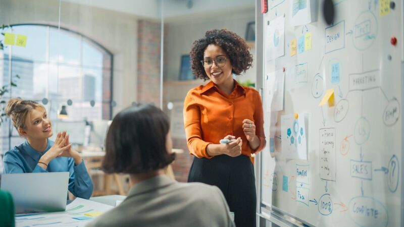 Meeting in the training room: a young, dark-skinned woman presents current facts and figures to two 