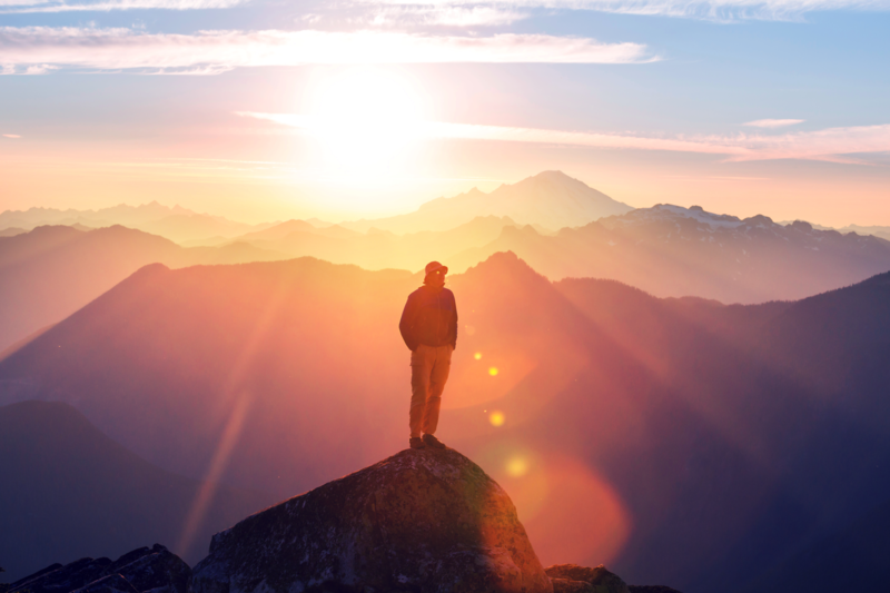 man in the mountains standing on a summit with the sun behind him