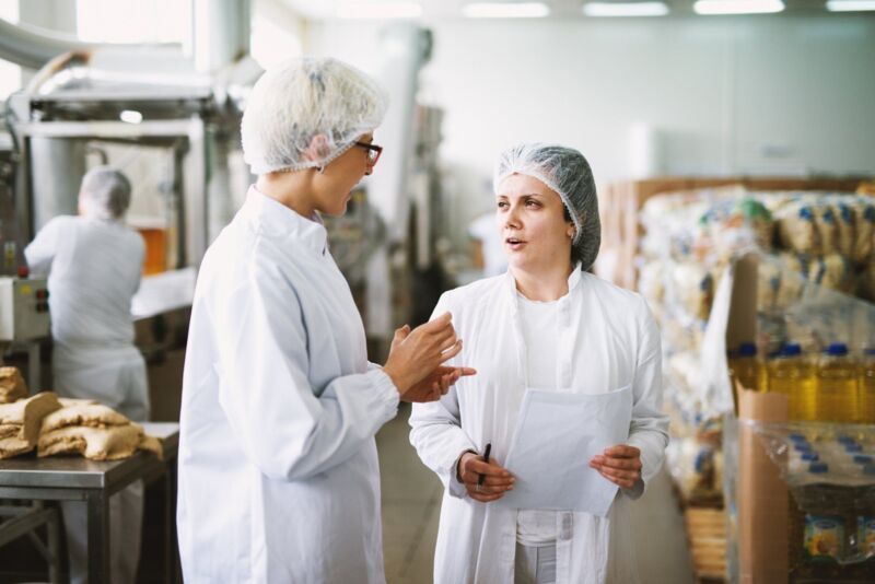Two cheerful workers in white sterile cloths are standing in a food factory and talking while one of