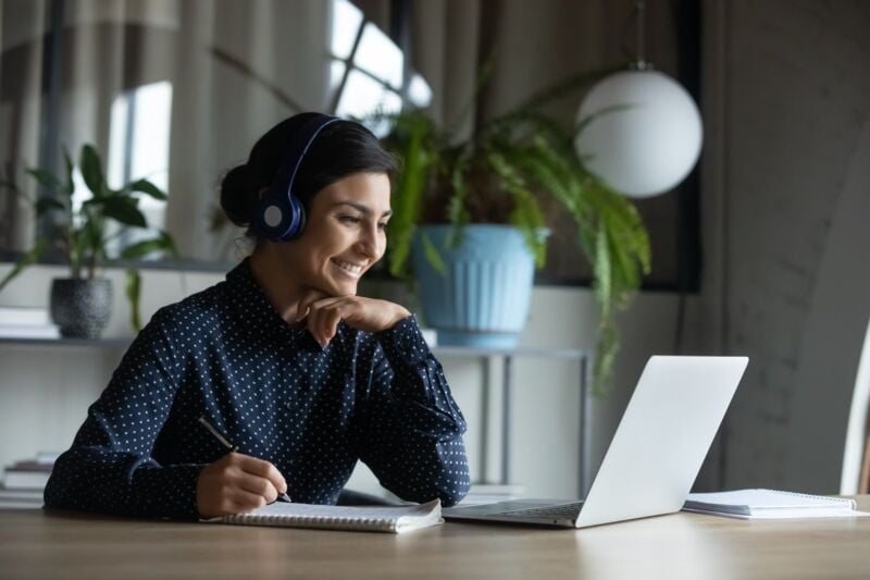 Happy young indian girl with wireless headphones looking at laptop screen, reading listening online 