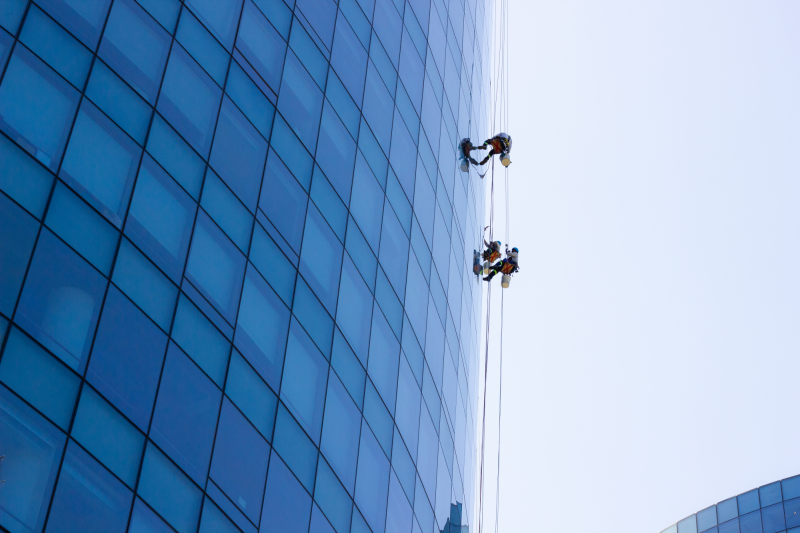 three window cleaners hang high up on a blue glass façade of an office building and clean the panes