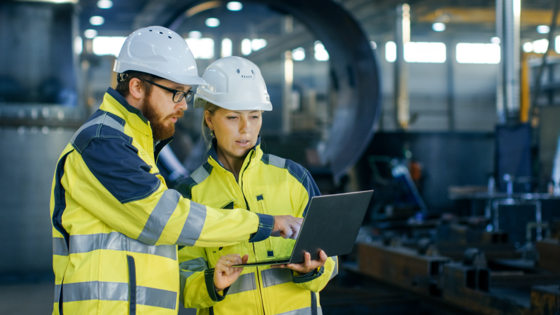 Male and Female Industrial Engineers in Hard Hats Discuss New Project while Using Laptop. They Make 